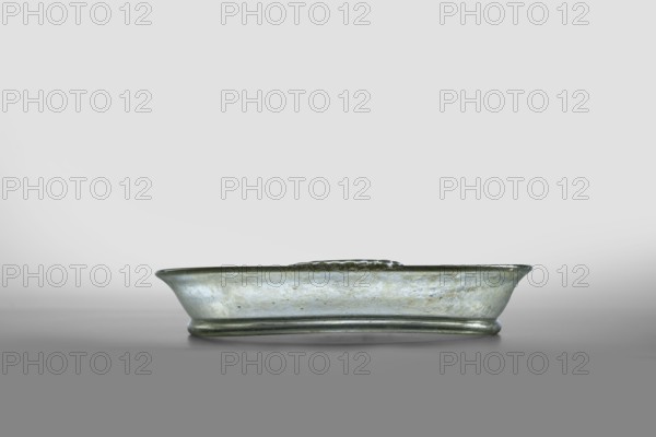 Plate, unknown eastern workshop; horizontal handles with ribs added to the rim edge; eastern workshop; Rome; late 1st–2nd century 1st–2nd century; Roman Empire 27 BC–476. Type: Isings form 43. Transparent, colorless glass with light green shade; free-blown. Outer diameter of rim: 14.50 cm; height: 1.90 cm; outer diameter of bottom: 12 cm; weight: 0.134 kg., A shallow, oblong glass dish with a slightly flared rim sits centered on a smooth surface; the glass shows a muted greenish-gray hue with areas of pale bluish and silvery sheen, and the surface has subtle mottled and weathered textures.