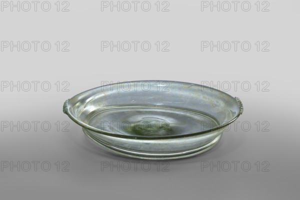 Plate, unknown eastern workshop; horizontal handles with ribs added to the rim edge; eastern workshop; Rome; 1st–2nd century late 1st–2nd century; Roman Empire 27 BC–476; Isings form 43; transparent, colorless glass with a light green shade; free-blown; outer rim diameter 14.50 cm; height 1.90 cm; outer bottom diameter 12 cm; vessel; weight 0.134 kg., A shallow round glass dish with a rim and a slightly raised base, shown from an angled top view; the dish appears translucent with greenish and gray tones and shows areas of subtle iridescence and surface imperfections; there are a few decorative ridges on the outer side and small ornamental details on opposite sides of the rim; the background is a smooth gradient in shades of light gray and darker gray at the bottom.