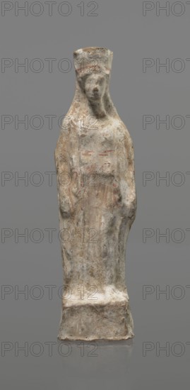 Female terracotta figurine of a standing woman, depicted frontally. Set on a small hollow, bottomless base rectangular at the front and oval at the back; a large rectangular opening at the back shows clear traces of carving in the unfired clay. The woman stands with legs slightly apart, weight on the straight right leg, left leg bent and slightly forward. Arms lowered, slightly bent at the elbows, hands holding a cloak at hip level. Head turned slightly to the left on a long neck; elongated face with very faint features; long combed-back hair over the shoulders. Polos on her head decorated with a horizontal red line. Garments fit snugly with plastically defined folds. Traces of red polychrome on a white ground cover the entire surface; lines highlight garment folds and mark a necklace and the belt tying the peplos. Damaged polychrome. 5th century BC, Greece. Height 19.6 cm., A weathered standing figure sculpture made of a light-colored stone with areas of brown and reddish discoloratio...