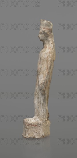 Female terracotta figurine of a standing woman, depicted frontally. Set on a small hollow, bottomless base, rectangular at the front and oval at the back. A large rectangular opening at the back shows clear traces of carving in the unfired clay. The woman stands with legs slightly apart, weight on the straight right leg, left leg bent and slightly forward. Arms lowered, slightly bent at the elbows, hands holding a cloak at hip level. Head turned slightly to the left on a long neck; elongated face with very faint features. Long combed-back hair flows over her shoulders. A polos on her head is decorated with a horizontal red line. Garments fit snugly with plastically defined folds. Traces of red polychrome on a white ground cover the entire surface; lines highlight garment folds and mark a necklace, and the belt tying the peplos. Damaged polychrome. 5th century BC, Greece. Height 19.6 cm., A weathered standing figure sculpture on a rounded rectangular base, shown in profile facing left, ...