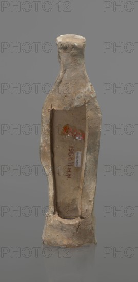 Female terracotta figurine of a standing woman, depicted frontally. Set on a small hollow bottomless base, rectangular at the front and oval at the back. A large rectangular opening at the back shows clear traces of carving in the unfired clay. The woman stands with legs slightly apart, weight on the straight right leg, left leg bent slightly forward. Arms lowered, slightly bent at the elbows, hands holding a cloak at hip level. Head turned slightly to the left on a long neck; elongated face with very faint features. Long combed-back hair flows over her shoulders. A polos on her head is decorated with a horizontal red line. Garments fit snugly with plastically defined folds. Traces of red polychrome on a white ground cover the entire surface; lines highlight the folds of the garment and mark a necklace, and the belt tying the peplos. Damaged polychrome. 5th century BC, Greece. Height 19.6 cm., A narrow, upright ceramic or clay vessel with rough, weathered surface and an open hollow fro...