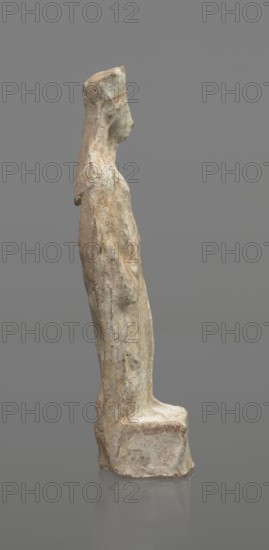 Female terracotta figurine of a standing woman, depicted frontally. Set on a small, hollow, bottomless base, rectangular at the front and oval at the back. A large, rectangular opening at the back shows clear traces of carving in the unfired clay. The woman stands with legs slightly apart, weight on the straight right leg; left leg bent and slightly forward. Arms lowered, slightly bent at the elbows, hands holding a cloak at hip level. Head turned slightly to the left on a long neck; elongated face with very faint features. Long, combed-back hair over the shoulders. Wears a polos decorated with a horizontal red line. Garments fit snugly with plastically defined folds. Traces of red polychrome on a white ground cover the entire surface; lines highlight the garment folds and mark a necklace, and the belt tying the peplos. Damaged polychrome. 5th century BC, Greece. Height 19.6 cm., A standing carved figure viewed in profile on a plain gray background, rendered in weathered pale stone ton...