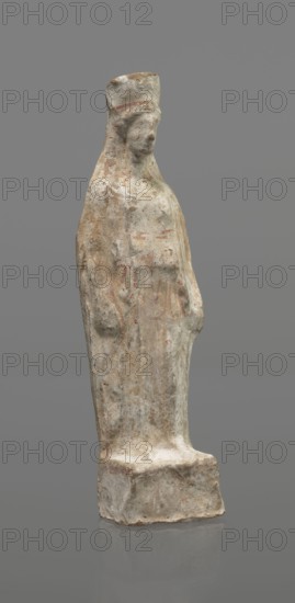 Female figurine, terracotta, figurine of a standing woman depicted frontally. Set on a small, hollow, bottomless base rectangular at the front and oval at the back. A large rectangular opening at the back shows clear traces of carving in the unfired clay. The woman stands with legs slightly apart, weight on the right leg; left leg bent slightly forward. Arms lowered, slightly bent at the elbows, hands hold a cloak at hip level. Head turned slightly to the left on a long neck; elongated face with very faint features. Long, combed-back hair flows over her shoulders. A polos on her head is decorated with a horizontal red line. Garments fit snugly with plastically defined folds. Traces of red polychrome on a white ground cover the entire surface; lines highlight garment folds and mark a necklace and the belt tying the peplos. Damaged polychrome. 5th century BC, Greece. Height 19.6 cm., A weathered standing figure carved from a pale stone with a rough, textured surface, showing areas of bro...