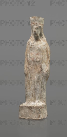 Female terracotta figurine of a standing woman, depicted frontally. Set on a small, hollow, bottomless base, rectangular at the front and oval at the back. A large, rectangular opening at the back shows clear traces of carving in the unfired clay. The woman stands with legs slightly apart, weight on the straight right leg, left leg bent slightly forward. Arms lowered, slightly bent at the elbows; hands hold a cloak at hip level. Head turned slightly to the left on a long neck; elongated face with very faint features; long combed-back hair over the shoulders. A polos on her head is decorated with a horizontal red line. Garments fit snugly with plastically defined folds. Traces of red polychrome on a white ground cover the entire surface; lines highlight garment folds and mark a necklace, and the belt tying the peplos. Damaged polychrome. 5th century BC, Greece. Height 19.6 cm., A standing sculpted figure on a rectangular base, rendered in weathered off-white and beige tones with areas o...