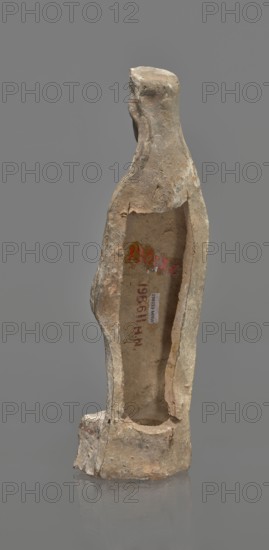 Female terracotta figurine of a standing woman, depicted frontally. Set on a small, hollow, bottomless base, rectangular at the front and oval at the back. A large rectangular opening at the back shows clear traces of carving in the unfired clay. The woman stands with legs slightly apart, weight on the right leg, left leg bent and slightly forward. Arms lowered, slightly bent at the elbows, hands holding a cloak at hip level. Head turned slightly left on a long neck; elongated face with very faint features. Long, combed-back hair flows over the shoulders. A polos on her head is decorated with a horizontal red line. Garments fit snugly with plastically defined folds. Traces of red polychrome on a white ground cover the entire surface; lines highlight garment folds and mark a necklace, and the belt tying the peplos. Damaged polychrome. 5th century BC, Greece. Height 19.6 cm., A weathered, narrow vertical sculpture with an elongated neck and simplified head stands against a neutral gray b...