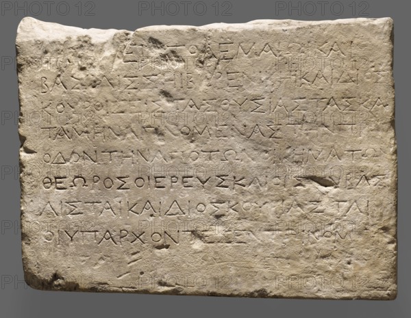 Plate with a Greek dedication to Ptolemy III, Berenice and the Dioscuri;, Greek inscriptions; Ptolemy III Euergetes ruler of Egypt, 246–221 BC; dedications; dated 246–241 BC before 241 BC, no epithet theoi Euergetai; Egypt; height 30.2 cm, width 40.5 cm, depth 7.7 cm, weight 15.728 kg, A rectangular stone slab with an uneven, worn surface and rounded corners rests against a neutral gray background; the slab is light beige with areas of darker beige and pale gray discoloration, and it shows surface pitting, chips, and shallow grooves across its face. The top edge is irregular and slightly raised in places, and there are a few shallow depressions and rough patches scattered across the surface. The overall texture appears weathered and slightly grainy, with subtle shadows emphasizing the carved indentations and surface irregularities.