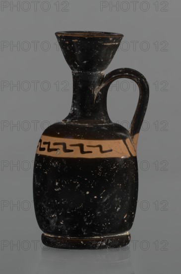 Black-finned aryballois lekythos, /workshop., repossession Silesia. Lekythos, black-finned pottery. Large, ring-shaped foot. Body squat, barrel-shaped with rounded shoulders. Neck narrow, clearly separated from body, with flaring rim. Upper part of body has a slightly irregular clay-colored orange band not covered with finnis; on it, at the front, a rather carelessly executed meander interrupted with black paint. Upper part of the vice is tilted to the left. Condition: minor chips, rim edge worn. Date: 1st half of the 19th century; 4th century BC. Unknown site/place of origin. Vessel lekythos. Height 10.9 cm; diameter 5.4 cm., A dark ceramic vessel with a narrow flared neck and a rounded body, featuring a single curved handle attaching from the upper neck to the shoulder; the surface shows worn spots and flecks revealing lighter underlying material, and a horizontal band of lighter color encircles the shoulder with a dark zigzag motif on it; visible colors include black, a tan or light...