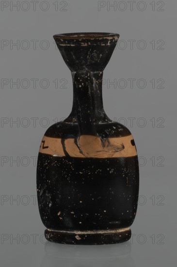 Black-finned aryballois lekythos, /workshop. Repossession Silesia. Lekythos in black-finned pottery. Large, ring-shaped foot. Body rather squat, barrel-shaped with rounded shoulders. Neck quite narrow, clearly separated from the body, and a flaring rim. Upper part of the body has a slightly irregular clay-colored orange band not covered with finnis; on it, at the front, a rather carelessly executed meander interrupted with black paint. Upper part of the vice is tilted to the left. Condition: minor chips, rim edge worn. 1st half of the 19th century; 4th century BC. Unknown place of origin. Vessel lekythos. Height 10.9 cm, diameter 5.4 cm., A single ceramic vessel photographed against a neutral gray background, featuring a narrow flared rim atop a tall neck that connects to a squatter rounded body and a low circular base; the surface shows a worn black finish with a horizontal band of tan clay around the upper body and scattered small lighter speckles and chips across the black areas, an...