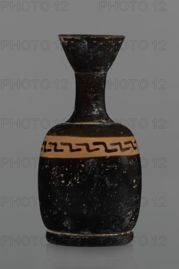 Black-finned aryballois lekythos. Large, ring-shaped foot. Body squat, barrel-shaped with rounded shoulders. Neck quite narrow, clearly separated from body with flaring rim. Upper part of body has a slightly irregular, clay-colored orange band not covered with finnis; on its front a rather carelessly executed meander interrupted with black paint. Upper part of the vice is tilted to the left. Condition: minor chips, rim edge worn. 1st half of the 19th century; 4th century BC., and place of origin. Vessel lekythos. Height 10.9 cm; diameter 5.4 cm., A small bottle-shaped ceramic vessel with a narrow neck flaring into a wide, shallow rim, showing a worn black surface with scattered lighter chips and speckles; the body has a horizontal band in a tan color around its upper portion decorated with a repeating wavy motif in black; the base is slightly raised and shows exposed areas where the black finish is worn to a lighter color; overall colors visible are black, tan, and small areas of light...