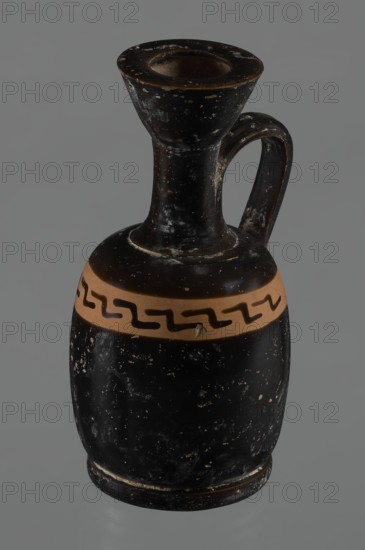 Black-finned aryballois lekythos, /workshop. Lekythos in black-finned pottery. Large ring-shaped foot; body squat, barrel-shaped with rounded shoulders. Narrow neck clearly separated from body and flaring rim. Upper part of body has a slightly irregular, clay-colored orange band not covered with finish; on it, at the front, a rather carelessly executed meander interrupted with black paint. Upper part of the vice is tilted to the left. Condition: minor chips, rim edge worn. Dated 4th century BC 1st half of the 19th century provenance note. Vessel lekythos. Height 10.9 cm; diameter 5.4 cm., A ceramic vessel with a narrow flared mouth, a tall neck, a curved handle attached near the rim, and a rounded body, showing surface wear and small chips; the vessel is primarily black with a band of tan around the upper body and a dark wavy pattern on that band, and the background is gray.