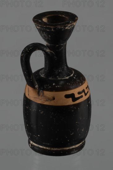 Black-finned aryballois lekythos, /workshop., repossession Silesia. Lekythos, black-finned pottery. Large, ring-shaped foot. Body squat, barrel-shaped with rounded shoulders. Neck quite narrow, clearly separated from body and flaring rim. Upper part of body has a slightly irregular, clay-colored orange band not covered with finish; on it, front of vessel, a rather carelessly executed meander interrupted with black paint. Upper part of the vice is tilted to the left. Condition: minor chips, rim edge worn. 1st half of the 19th century; 4th century BC. Unknown place of origin. Vessel, lekythos; height 10.9 cm, diameter 5.4 cm., A small ceramic vessel with a narrow neck, flared rim, and a curved handle attached between the neck and shoulder; the body is rounded and rests on a slightly raised base. The surface shows worn and chipped areas with a glossy black finish over most of the vessel and a band of natural clay color around the shoulder that includes a few abstract painted shapes in bla...