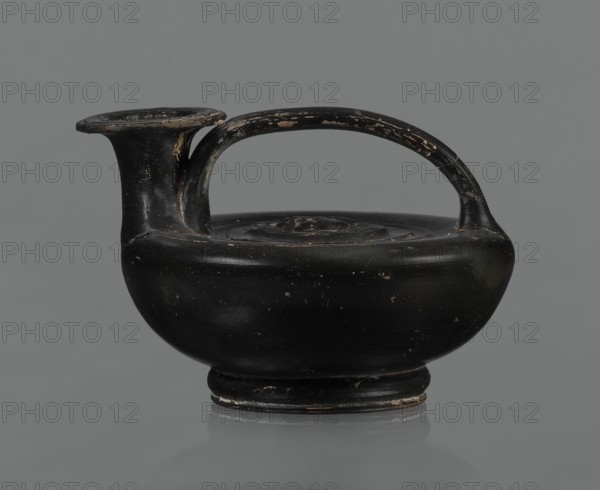 Black-finished askos, unknown Campanian workshop, black-finished pottery, askos, Campania Italy. Askos with a profiled foot and a semicircular body closed at the top with a slightly recessed surface. Narrow neck ending in a wide rim. Ribbon-shaped handle attached to the neck and to the upper surface of the vessel. Appliqué with a female head glued to the upper surface of the vessel, probably Medusa, surrounded by a wreath of intertwined snakes. Finish is black, shiny, worn in places, particularly on the handle and the edge of the rim. Light orange clay. Handle and neck cracked and glued. Mid 3rd century BCE., A rounded ceramic vessel with a single arched handle and a short spout, shown against a plain gray background; the surface of the vessel is predominantly black with areas of worn paint revealing lighter, tan-colored spots and edges, and there are small scuffs and chips across its body and handle.