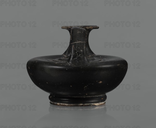 Black-finished askos, unknown Campanian workshop, black-finished pottery, askos, Campania Italy. Askos with a profiled foot and a semicircular body closed at the top with a slightly recessed surface. Narrow neck ending in a wide rim. Ribbon-shaped handle attached to the neck and to the upper surface of the vessel. Appliqué with a female head on the upper surface, probably Medusa, surrounded by a wreath of intertwined snakes. Finish is black, shiny, worn in places, particularly on the handle and the rim edge. Light orange clay. Handle and neck cracked and glued. Mid 3rd century BC., A black rounded ceramic vessel with a short neck and a flat, T-shaped rim sits centered against a neutral gray background; the surface shows worn areas with small chips and lighter scratches revealing a tan or brownish underlayer, and the base rim has patches of lighter wear.