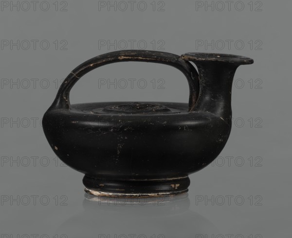 Black-finished askos, unknown Campanian workshop, repossession Silesia, black-finished pottery, askos, Campania Italy. Askos with a profiled foot and a semicircular body closed at the top with a slightly recessed surface. Narrow neck ending in a wide rim. Ribbon-shaped handle attached to the neck and to the upper surface of the vessel. Appliqué with a female head glued to the upper surface of the vessel, probably Medusa, surrounded by a wreath of intertwined snakes. Finish is black, shiny, worn in places, particularly on the handle and the edge of the rim. Light orange clay. Handle and neck cracked and glued. Mid 3rd century BC, Campania, vessel., A black ceramic vessel with a rounded body, a short pedestal base, and a wide flat rim connected to the body by a tall, flaring neck; a curved handle arches from one side of the rim to the opposite shoulder. The surface shows areas of wear where the black coating is chipped, revealing lighter clay beneath, and there are small scratches and sc...