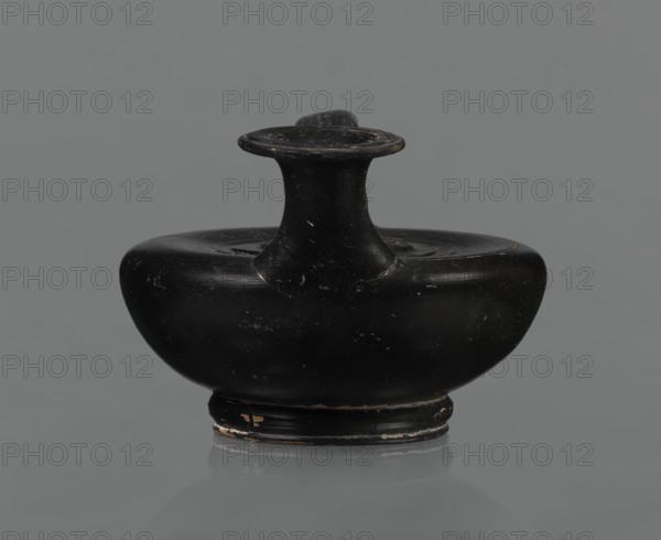 Black-finished askos, unknown Campanian workshop, black-finished pottery, askos, Campania Italy. Askos with a profiled foot and a semicircular body closed at the top with a slightly recessed surface. Narrow neck ending in a wide rim. Ribbon-shaped handle attached to the neck and to the upper surface of the vessel. Appliqué of a female head glued to the upper surface of the vessel, probably Medusa, surrounded by a wreath of intertwined snakes. Finish is black, shiny, worn in places, particularly on the handle and the edge of the rim. Light orange clay. Handle and neck cracked and glued. Mid 3rd century BC., A small rounded ceramic vessel with a wide flattened body and a short flared neck topped by a flat disc-like rim, shown against a plain neutral background; the surface is predominantly black with visible small areas of worn lighter color and slight scuffs, and the base shows a few patches of lighter wear; the overall shape is symmetrical and compact with smooth curved contours and a ...