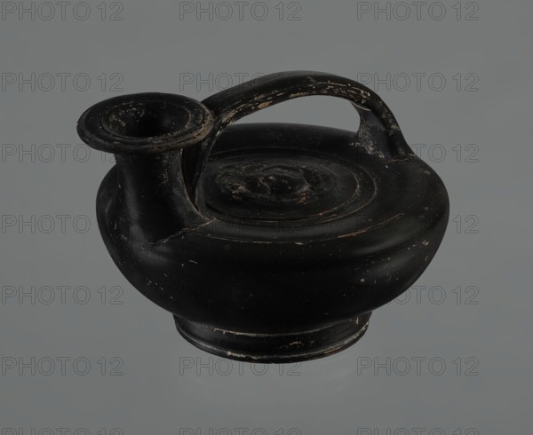 Black-finished askos, unknown Campanian workshop, repossession Silesia, black-finished pottery, askos, Campania Italy, Askos with a profiled foot and a semicircular body closed at the top with a slightly recessed surface. Narrow neck ending in a wide rim. Ribbon-shaped handle, attached to the neck and to the upper surface of the vessel. Appliqué with a female head glued to the upper surface of the vessel. The head depicted is probably Medusa, surrounded by a wreath of intertwined snakes. The finish is black, shiny, worn in places, particularly on the handle and the edge of the rim. Light orange clay. Handle and neck cracked and glued. Mid. III wa C., Campania, vessel, A black rounded ceramic vessel with a short flared spout and an arched handle, showing worn and chipped areas that reveal lighter tan and off-white underlying material, resting against a uniform gray background; the vessel's surface has subtle circular ridges and scuffs with a few small lighter flecks.