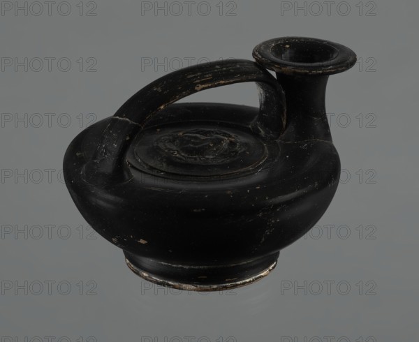 Black-finished askos, unknown Campanian workshop, black-finished pottery, askos, Campania Italy, Askos with a profiled foot and a semicircular body closed at the top with a slightly recessed surface. Narrow neck ending in a wide rim. Ribbon-shaped handle attached to the neck and to the upper surface of the vessel. Appliqué with a female head glued to the upper surface of the vessel. The head depicted is probably Medusa, surrounded by a wreath of intertwined snakes. The finish is black, shiny, worn in places, particularly on the handle and the edge of the rim. Light orange clay. Handle and neck cracked and glued. Mid 3rd century BCE, Campania, vessel., A black rounded ceramic vessel with a single curved handle arching over the top and a short flared spout, showing worn areas with small patches of lighter color and surface scratches, sitting against a plain gray background.
