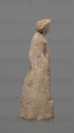 Terracotta figurine of Heracles. Figurine of a standing naked Heracles in a contrapposto pose with a cloak thrown over his back. His right hand rests on a rock, and in his left he probably holds a club. Terracotta, made from a mold. Figurine of a nude male, Greek mythology. Condition: good. Date: 2nd–1st century BC. Place of origin: Asia Minor. Height: 14.3 cm., A roughly carved standing figure of a human form made of a light tan, beige material with areas of darker brown and faint yellowish spots, showing a simplified head, shoulders, and torso; the surface is textured with visible pitting and small cracks, and there are worn, smoother areas on the torso and head. The figure is viewed in profile, with one arm suggested against the body and the lower portion flaring into a skirt-like shape; the base is uneven and rests on a smooth reflective surface. The background is a uniform medium gray.
