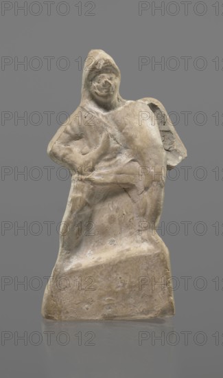 Terracotta gladiator figurine, unknown Southern Italian workshop, terracotta, male figurine. Figure of a gladiator ready for battle on a high base. Legs wide apart, left leg bent, right leg straight. On the calves are greaves reaching above the knees. In his right, bent hand he holds a dagger, and in the other a four-sided shield. He is dressed in a short draped tunic and wears a helmet. The Latin inscription PACILI is visible on the reverse of the base. Preservation condition: part of the base filled with plaster, partial shield and back of the head broken off, reflections, cracks. 2nd–1st century BC, Southern Italy, figurine; height 13 cm, width 7 cm., A weathered stone figurine of a human-like figure standing on a rectangular base, shown against a solid gray background; the figure wears a hood and has a rounded head with faint facial features, a sloped shoulder and an arm bent across the torso, and a curved object or draped element over one shoulder; the surface is rough and pitted ...