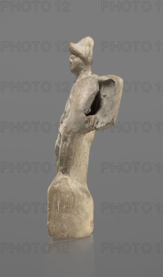 Terracotta gladiator figurine, unknown Southern Italian workshop, terracotta, male figurines. Figure of a gladiator ready for battle on a high base. Legs wide apart, left leg bent, right leg straight. On the calves are greaves reaching above the knees. In his right, bent hand he holds a dagger, and in the other a four-sided shield. He is dressed in a short draped tunic and wears a helmet. The Latin inscription PACILI is visible on the reverse of the base. Preservation condition: part of the base filled with plaster, partial shield and back of the head broken off, reflections, cracks. Date: 2nd–1st century BC, Southern Italy. Figurine; height 13 cm, width 7 cm., A small sculpted figure made of a pale beige material stands against a solid gray background. The figure appears to wear a hat and has a simplified, abstracted form with a rounded head, a narrow torso, and elongated limbs. A curved element projects from the figure's back like a flattened, hollowed shape. The surface shows rough ...