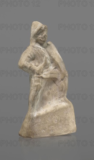 Terracotta gladiator figurine, unknown Southern Italian workshop, terracotta, male figurines. Figure of a gladiator ready for battle on a high base. Legs wide apart, left leg bent, right leg straight. Greaves reaching above the knees. In his right, bent hand he holds a dagger, and in the other a four-sided shield. Dressed in a short draped tunic and wearing a helmet. The Latin inscription PACILI is visible on the reverse of the base. Preservation: part of the base filled with plaster, partial shield and back of the head broken off, reflections, cracks. Date: 2nd–1st century BC. Place of origin: Southern Italy. Figurine, height 13 cm, width 7 cm., A small weathered sculpture of a standing figure carved from a pale beige material, showing a rough, uneven surface with worn details; the figure appears to wear a hood or hat and has indistinct facial features, with one arm bent and the other resting along the body; the lower portion broadens into a solid base with textured marks and chips; t...