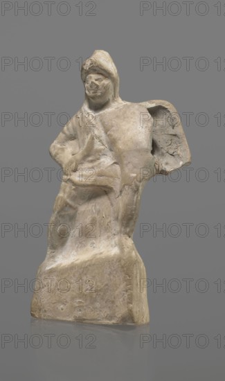Terracotta gladiator figurine, unknown Southern Italian workshop, terracotta, male figurines. Figure of a gladiator ready for battle on a high base. Legs wide apart, left leg bent, right leg straight. On the calves are greaves reaching above the knees. In his right, bent hand he holds a dagger, and in the other a four-sided shield. He is dressed in a short draped tunic and wears a helmet. The Latin inscription PACILI is visible on the reverse of the base. Preservation condition: part of the base filled with plaster, partial shield and back of the head broken off, reflections, cracks. Date: 2nd–1st century BC, Southern Italy. Figurine, height 13 cm, width 7 cm., A weathered, pale beige stone figurine of a seated person wearing a hooded garment is shown against a neutral gray background; the figure has softened, eroded facial features and simplified limbs, with a draped cloak that creates folds and rounded forms, and a rough, blocky base that the figure merges into. The object displays p...