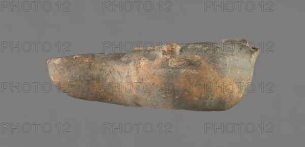 Oil lamp, unknown Gaulish workshop, repossession Frombork; Warmian-Masurian Voivodeship, oil lamps, single-burner lamp, Roman lamp, Buchi type IX-b = Loeschcke type IXb oil lamp. Lamp from a mold. Round vessel. Arms sloping outward, separated from the disc by a convex edge. Two bars arranged symmetrically on the arms. One vertically placed vice opposite the burner also from a mold, broken off. Concave, flat disc with one large centrally located olive hole. Medium-length wick channel, decorated with a groove in the center. Two small air ventilation holes at either end of the groove. Rounded end of the burner. Large wick hole. No charring. 1st century, Gaul, oil lamp; height 3.7 cm, width 10.9 cm, depth 7 cm., An elongated, weathered object with an irregular, rounded form appears against a plain gray background, showing a textured surface with a mix of muted brown, tan, ochre, and gray tones; the surface has rough, pitted areas and smoother patches, with subtle variations in color and sc...