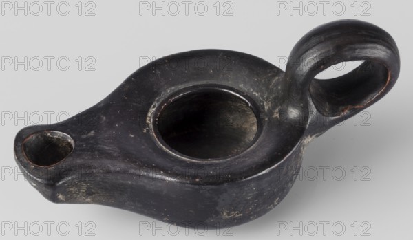 Oil lamp, unknown Attic workshop; oil lamp, single-burner, lighting; scene: gallery/classical; end of 4th century BC; Attica; weight 0.090 kg., A dark, rounded object with a smooth, worn surface and a glossy finish, showing areas of lighter wear and small patches of rough texture; it has a central open cavity, a smaller hollow spout on one side, and a large looped handle on the other, with curved, flowing contours and visible interior hollows; the visible colors are predominantly black and dark gray with spots of lighter gray and hints of brownish tones along some edges.