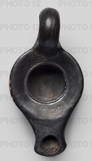 Oil lamp, unknown Attic workshop; oil lamp, single-burner, lighting; scene: gallery/classical?; end of 4th century BC; Attica; weight 0.090 kg., A dark, rounded object with a smooth, worn surface and a glossy sheen, primarily black with areas of brownish wear and small patches of lighter tan showing through; it has a raised, curved handle at one end and a circular recessed basin near the center with visible concentric surface marks, and a smaller rounded opening at the opposite end with a slightly lighter interior; the background is a pale gray.
