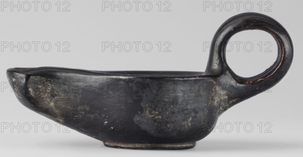 Oil lamp, unknown Attic workshop; oil lamp, single-burner lamp, lighting; scene gallery class; end of 4th century BC; Attica; weight 0.090 kg, A low, shallow vessel with an elongated, spouted front and a rounded rear that transitions into a large circular handle; the surface is predominantly dark black with areas of muted gray and worn lighter patches showing irregular mottling; subtle brownish and rusty tones appear along worn edges and the handle interior; the piece sits on a small foot and shows a slightly glossy, uneven finish with visible texture and wear.