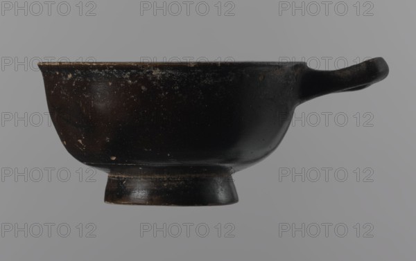 Black-finned goblet, unknown Attic workshop, repossession Zabkowice Slaskie, ceramics, black-finned pottery, Attica Greece, Greek pottery, bowls vessels, antiquity, Ancient Greece, ancient vessels. Black-finned goblet with a deep bowl with a wide, slightly outward-turning rim, one cylindrical handle attached horizontally just above the rim, and a low, ring-shaped foot. The glossy black finish covers the entire vessel except for the inside of the foot. 4th century BC, Attica, vessel, goblet, height 4.5 cm, width 8.5 cm, A dark brown rounded vessel with a short pedestal base and a small handle projecting from one side, shown against a gray background; the surface of the vessel has a worn, slightly rough texture with small lighter scuffs and spots, and there are subtle variations of dark brown and black on the body and handle.