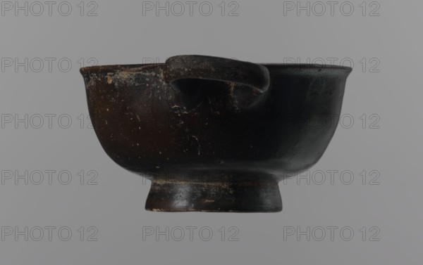 Black-finned goblet, unknown Attic workshop, black-finned pottery, Attica Greece, Greek pottery, bowls vessels, antiquity, Ancient Greece, ancient vessels. Black-finned goblet with a deep bowl with a wide, slightly outward-turning rim, one cylindrical handle attached horizontally just above the rim, and a low, ring-shaped foot. The glossy black finish covers the entire vessel except for the inside of the foot. 4th century BC, Attica, vessel, goblet, height 4.5 cm, width 8.5 cm., A dark bowl with a rounded body and a short pedestal base, showing worn and chipped areas on its surface and rim, with a small loop handle attached near the top; the bowl appears mostly black with hints of brown in worn spots and lighter speckles, set against a neutral gray background.
