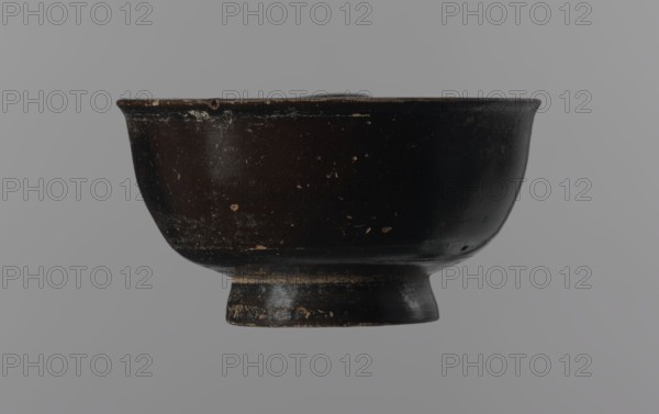 Black-finned goblet, unknown Attic workshop, repossession Zabkowice Slaskie, ceramics, black-finned pottery, Attica Greece, Greek pottery, bowls vessels, antiquity, Ancient Greece, ancient vessels. Black-finned goblet with a deep bowl with a wide, slightly outward-turning rim, one cylindrical handle attached horizontally just above the rim, and a low, ring-shaped foot. The glossy black finish covers the entire vessel except for the inside of the foot. 4th century BC, Attica, vessel, goblet, height 4.5 cm, width 8.5 cm., A dark bowl with a rounded body and a short pedestal base sits against a plain light gray background; the bowl's surface is predominantly black with worn areas showing brown and beige flecks and scuffs, and the rim and base show uneven wear and small chips.