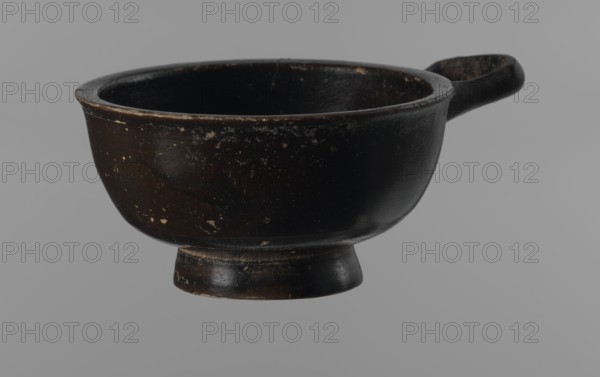 Black-finned goblet, unknown Attic workshop, black-finned pottery, Attica Greece, Greek pottery, bowl-shaped vessel, antiquity, Ancient Greece. Black-finned goblet with a deep bowl with a wide, slightly outward-turning rim, one cylindrical handle attached horizontally just above the rim, and a low, ring-shaped foot. The glossy black finish covers the entire vessel except for the inside of the foot. 4th century BC. Vessel/goblet. Height 4.5 cm; width 8.5 cm., A dark brown bowl with a short pedestal base and a small handle projecting from one side, the bowl showing worn areas and small lighter speckles on its exterior and interior, photographed against a plain light gray background; visible colors include dark brown, black, lighter brown spots, and gray.