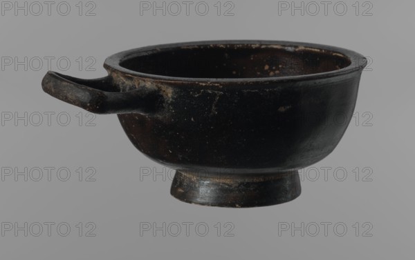 Black-finned goblet, unknown Attic workshop, repossession Zabkowice Slaskie, ceramics, black-finned pottery, Attica Greece, Greek pottery, bowls vessels, antiquity, Ancient Greece, ancient vessels. Black-finned goblet with a deep bowl with a wide, slightly outward-turning rim, one cylindrical handle attached horizontally just above the rim, and a low, ring-shaped foot. The glossy black finish covers the entire vessel except for the inside of the foot. 4th century BC, Attica, vessel, goblet, height 4.5 cm, width 8.5 cm, A dark, shallow bowl with a short pedestal base and a small protruding handle, shown against a neutral gray background; the bowl appears worn with rough patches and speckled surface texture, and visible colors include black, dark brown, and areas of lighter brown or tan where the surface is rubbed.