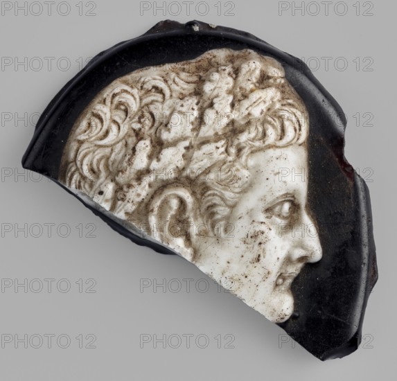 Cameo with a representation of the head of Tiberius in an oak wreath, unknown Italian workshop; male head iconography; glass; Tiberius Roman emperor; 14–37; gems; ca. 20; Italy, Roman Empire 27 BC–476; cameo; height 3.5 cm, width 3.5 cm, depth 0.8 cm, weight 0.008 kg., A fragment of a carved relief showing a right-facing human profile with curly hair and a laurel wreath, rendered in off-white with areas of brownish dirt or staining, set against a dark glossy black background with irregular broken edges; the overall scene includes shades of gray in the surrounding backdrop.