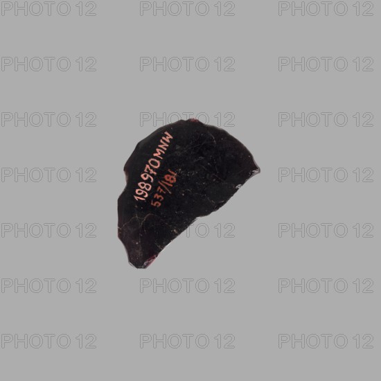 Cameo with a representation of the head of Tiberius in an oak wreath, unknown Italian workshop; male head iconography; glass; Tiberius Roman emperor; 14–37; gems; ca. 20; Italy, Roman Empire 27 BC–476; cameo; height 3.5 cm, width 3.5 cm, depth 0.8 cm, weight 0.008 kg., A dark, irregularly shaped fragment with a glossy black surface and rough, chipped edges sits against a light gray background; the fragment shows a few small areas of deep red along its edges and some pale scratch-like marks on its surface, and there is a short vertical grouping of colored markings on the fragment.
