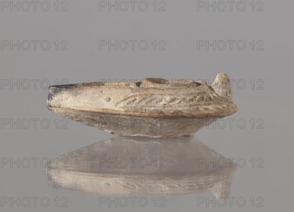 Oil lamp, unknown Palestinian workshop. Oval lamp with conical handle. Large olive opening occupying virtually the entire surface of the disc. Arms decorated with branches. Burner channel and wick opening bounded by a convex shaft. The channel decorated with a six-leafed palm branch, interpreted in the context of Byzantine Jerusalem workshops as a schematic representation of a menorah. Oval, ring-shaped base decorated in the center with a six-pointed star Magen of David, known in Arab-Muslim culture as the Seal of Solomon. On the outside of the ring-shaped base are three symmetrically spread palm branches—menorahs. Entire lamp impressed into the mold. Islamic art; Arabic culture; oil lamp used in everyday life in the Middle East; iconography: palm branches, star, Seal of Solomon, six-pointed star. 7th–8th century, Syro-Palestine. Dimensions: height 3.4 cm; length 10.8 cm; width 6.9 cm., A small shallow vessel with a rounded base and an upturned rim sits on a reflective surface, showing...