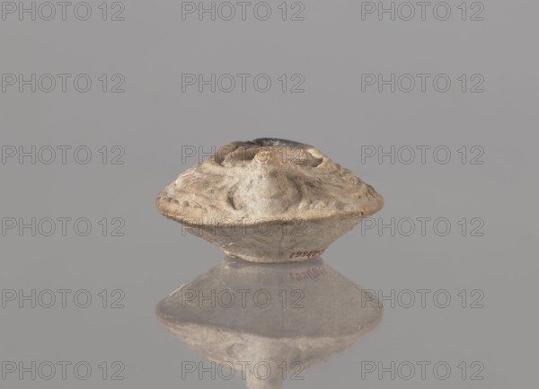 Oil lamp, unknown Palestinian workshop. Oval lamp with conical handle. Large olive opening occupying virtually the entire surface of the disc. Arms decorated with branches. Burner channel and wick opening bounded by a convex shaft. Channel decorated with a six-leafed palm branch, interpreted in the context of Byzantine Jerusalem workshops as a schematic representation of a menorah. Oval, ring-shaped base decorated in the center with a six-pointed star Magen David / Seal of Solomon. Outside the ring-shaped base are three symmetrically spread palm branches—menorahs. Entire lamp impressed from a mold. Islamic art, Arabic culture; 7th–8th century, Syro-Palestine. Oil lamp; height 3.4 cm, length 10.8 cm, width 6.9 cm., A small, weathered object with an irregular, rounded shape sits centered against a smooth, light gray background, showing a textured surface in shades of beige, tan, and light brown with some darker brown spots and a slightly darker top area; the object casts a soft reflectio...