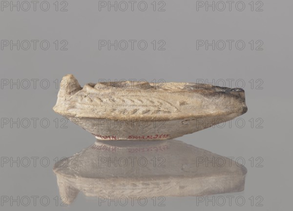 Oil lamp, unknown Palestinian workshop. Oval lamp with conical handle. Large olive opening occupying almost entire disc. Arms decorated with branches. Burner channel and wick opening bounded by a convex shaft; channel decorated with a six-leafed palm branch interpreted in Byzantine Jerusalem workshops as a schematic menorah. Oval ring-shaped base decorated in the center with a six-pointed star Magen David / Seal of Solomon. Outside the ring-shaped base are three symmetrically spread palm branches menorahs. Entire lamp was mold-impressed. 7th–8th century, Syro-Palestine. Oil lamp; height 3.4 cm, length 10.8 cm, width 6.9 cm. Arabic; Islamic art; palm branches and star iconography., A small shallow dish-like object sits on a reflective surface, showing a worn, irregular rim and a raised spout-like projection at one edge; the object appears textured with carved or molded linear patterns along its side and uneven indentations on its top. The visible colors are beige, tan, light brown, gray...