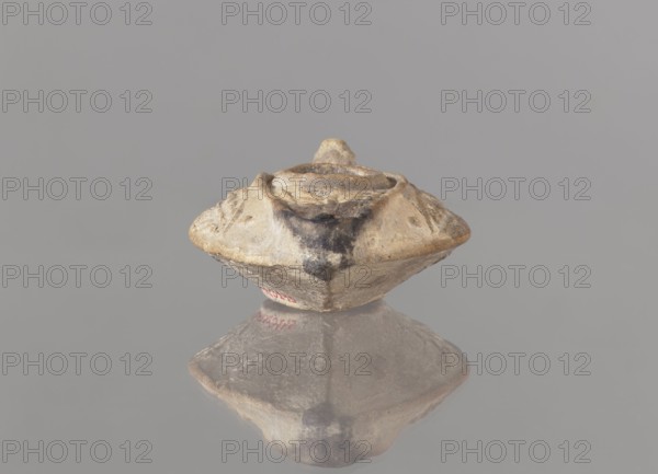 Oil lamp, unknown Palestinian workshop. Islamic art, Arabic culture. Oval lamp with conical handle. Large olive opening occupying virtually the entire surface of the disc. Arms decorated with branches. Burner channel and wick opening bounded by a convex shaft; channel decorated with a six-leafed palm branch, interpreted in the context of Byzantine Jerusalem workshops as a schematic representation of a menorah. Oval, ring-shaped base decorated in the center with a six-pointed star Magen David, known in Arab-Muslim culture as the Seal of Solomon. Outside the ring-shaped base are three symmetrically spread palm branches interpreted as menorahs. The entire lamp is impressed into the mold. 7th–8th century, Syro-Palestine. Oil lamp; height 3.4 cm, length 10.8 cm, width 6.9 cm., A small, worn object with an angular, rounded shape sits centered on a smooth reflective surface, showing a mirrored reflection beneath it; the object appears textured and uneven with rough edges and a shallow central...
