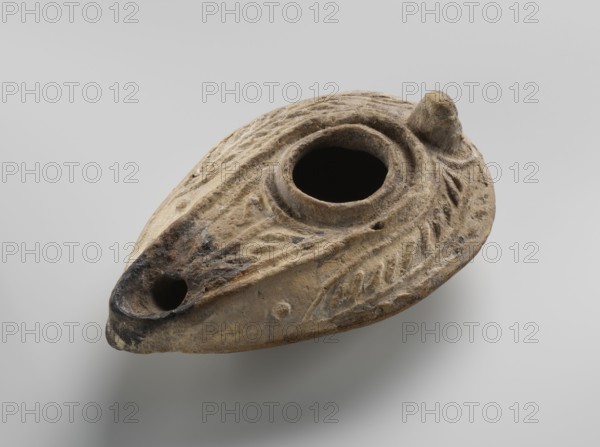 Oil lamp, unknown Palestinian workshop. Oval lamp with conical handle. Large olive opening occupying virtually the entire surface of the disc. Arms decorated with branches. Burner channel and wick opening bounded by a convex shaft; the channel decorated with a six-leafed palm branch, interpreted in the context of Byzantine Jerusalem workshops as a schematic representation of a menorah. Oval, ring-shaped base decorated in the center with a six-pointed star Magen David / Seal of Solomon. On the outside of the ring-shaped base are three symmetrically placed palm branches—menorahs. The entire lamp is impressed from a mold. 7th–8th century, Syro-Palestine. Islamic art; Arabic culture. Oil lamp, height 3.4 cm, length 10.8 cm, width 6.9 cm., An oval, shallow ceramic object with a central round opening and a tapered spout, showing textured carved patterns along its surface; the object appears worn with rough, pitted areas and a small knob-like protrusion near one edge. The visible colors inclu...