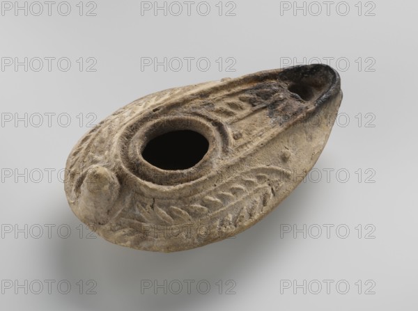 Oil lamp, unknown Palestinian workshop. Oval lamp with conical handle. Large olive opening occupying virtually the entire surface of the disc. Arms decorated with branches. Burner channel and wick opening bounded by a convex shaft. Channel decorated with a six-leafed palm branch, interpreted in the context of Byzantine Jerusalem workshops as a schematic representation of a menorah. Oval ring-shaped base decorated in the center with a six-pointed star Magen David, known in Arab-Muslim culture as the Seal of Solomon. Outside the ring-shaped base are three symmetrically spread palm branches—menorahs. Entire lamp impressed into the mold. 7th–8th century, Syro-Palestine. Materials/function: oil lamp. Dimensions: height 3.4 cm; length 10.8 cm; width 6.9 cm. Cultural/iconographic notes: Islamic art; Arabic; palm branches; star; Seal of Solomon; everyday life in the Middle East., A close-up of an oval, shallow clay object with a central round opening and a pointed spout at one end, showing a t...