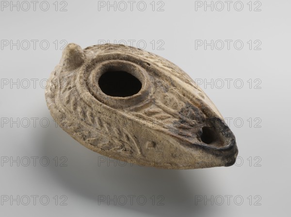 Oil lamp, unknown Palestinian workshop. Oval lamp with conical handle. Large olive opening occupying virtually the entire surface of the disc. Arms decorated with branches. Burner channel and wick opening bounded by a convex shaft. The channel decorated with a six-leafed palm branch, interpreted in the context of Byzantine Jerusalem workshops as a schematic representation of a menorah. Oval, ring-shaped base decorated in the center with a six-pointed star Magen David / Seal of Solomon. On the outside of the ring-shaped base are three symmetrically spread palm branches menorahs. Entire lamp impressed from a mold. 7th–8th century, Syro-Palestine. Oil lamp; height 3.4 cm, length 10.8 cm, width 6.9 cm., An oval, shallow object with a rough textured surface in shades of tan, beige, brown, and darker brown, featuring a large central circular opening and a smaller rounded opening at one tapered end, with carved or molded leaf-like patterns on the upper surface and a rounded knob near one edge...