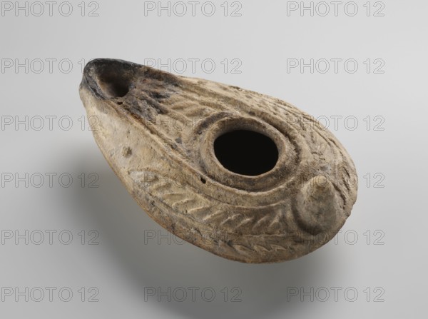 Oil lamp, unknown Palestinian workshop. Islamic art, Arabic culture; oil lamp used in everyday life in the Middle East. Oval lamp with conical handle. Large olive opening occupying virtually the entire surface of the disc. Arms decorated with palm branches. Burner channel and wick opening bounded by a convex shaft; the channel is decorated with a six-leafed palm branch, interpreted in the context of Byzantine Jerusalem workshops as a schematic representation of a menorah. Oval, ring-shaped base decorated in the center with a six-pointed star Magen David / Seal of Solomon. On the outside of the ring-shaped base are three symmetrically spread palm branches—menorahs. The entire lamp is impressed into the mold. 7th–8th century, Syro-Palestine. Oil lamp; height 3.4 cm, length 10.8 cm, width 6.9 cm., A worn, oval clay object with a central round opening and a smaller recess at one end, showing carved leaf-like patterns and raised ridges across its surface; the material appears rough and pitt...