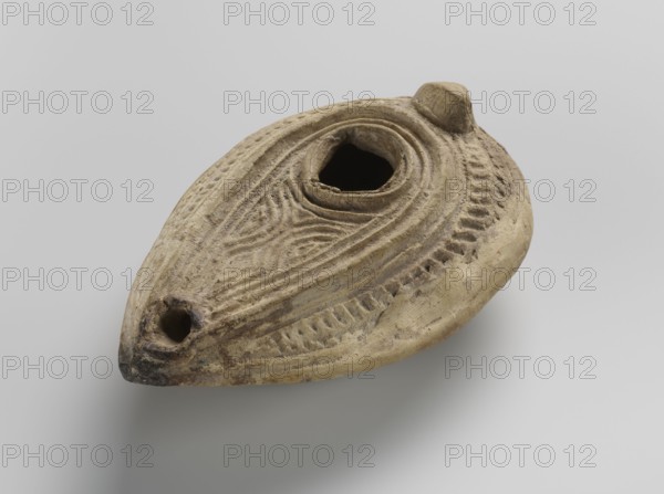 Oil lamp, Islamic art, Arabic culture; household ceramic oil lamp almond-shaped misbah zajti, modeled in two molds and glued together from yellow clay with impressed decorations; closed form with conical handle. Decorated on the upper surface with arched lines forming a palmette-shaped composition below the olive opening; sides of the arms decorated with fine lines. Inside the ring of the foot and on the bottom, the word baraka grace/blessing appears; remaining bottom surface decorated with chevron and grid patterns. Geometric ornament. Type A according to W. Kubiak's typology for al-Fustat dated 8th–10th century. Probable origin: Syria or Palestine. Dimensions: height 3.4 cm, length 10.7 cm, width 6.9 cm., A worn oval clay object with an irregular central opening and a pointed end with a small round hole, decorated with carved concentric and wavy grooves and ridges, showing rough texture and signs of wear; colors visible include shades of beige, tan, light brown, darker brown, and hin...