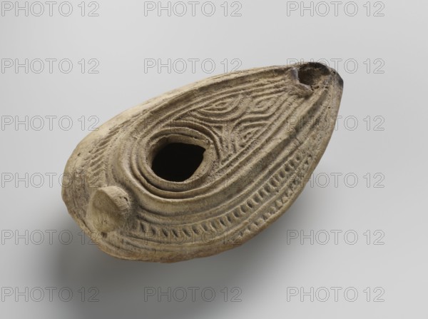 Oil lamp, Islamic art, Arabic culture, household ceramics, Arabic pottery, oil lamp, geometric ornament, Arabic inscriptions, everyday life in the Middle East. Decorated with geometric patterns over the surface inside the ring of the baraka foot. Yellow clay, modeled in two molds and glued together; impressed decorations. Almond-shaped lamp misbah zajti with a conical handle; closed form. Upper part decorated with arched lines forming a palmette-shaped composition below the olive opening; sides of the arms decorated with fine lines. On the bottom, within the foot, the word baraka grace, blessing, boon. Remaining surface of the bottom decorated with chevron and grid patterns. Classified as type A in W. Kubiak's typology for Al-Fustat M. Redlak. 8th–10th century. Possible origin: Syria or Palestine. Dimensions: height 3.4 cm, length 10.7 cm, width 6.9 cm., An oval, shallow carved object made of a beige to light brown material with a rough, weathered texture rests against a plain light gr...