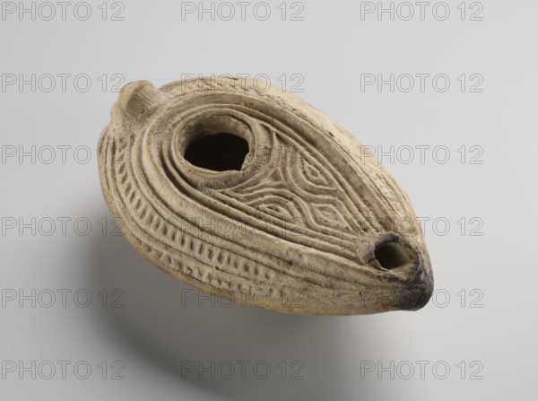 Oil lamp, Islamic art, Arabic culture; household ceramic oil lamp almond-shaped misbah zajti with conical handle, closed form; yellow clay modeled in two molds and glued together with impressed decorations. Decorated on the upper surface with arched lines forming a palmette-shaped composition below the olive opening; sides of the arms decorated with fine lines. Inside the ring of the foot is the Arabic inscription baraka “grace, blessing, boon”; remaining bottom surface decorated with chevron and grid patterns. Attributed to type A per W. Kubiak's typology for Al-Fustat; dated 8th–10th century; possible origin Syria or Palestine. Dimensions: height 3.4 cm, length 10.7 cm, width 6.9 cm., An oval, shallow ceramic object with a rounded protrusion on one end and a spout-like opening on the opposite end rests against a pale background; the surface is textured with carved concentric lines and geometric motifs, showing worn and rough areas, with visible shadows beneath it; the colors visible ...