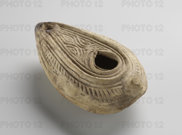 Oil lamp, Islamic art, Arabic culture, household ceramics, Arabic pottery, oil lamp, geometric ornament, Arabic inscriptions; decorated with geometric patterns over the entire surface, inside the ring of the baraka foot. Yellow clay, formed from two molds joined together; impressed decorations. Almond-shaped lamp misbah zajti with a conical handle; closed form. Upper part decorated with arched lines forming a palmette-shaped composition below the olive opening; sides of the arms decorated with fine lines. On the bottom, within the foot, appears the word baraka grace, blessing. Remaining surface of the bottom decorated with chevron and grid patterns. Classified as type A according to W. Kubiak's typology for Al-Fustat M. Redlak. Date: 8th–10th century. Possible origin: Syria or Palestine. Dimensions: height 3.4 cm; length 10.7 cm; width 6.9 cm., An oval, boat-shaped clay object in beige and light tan tones with darker brown-gray shading and weathered spots rests against a pale gray back...