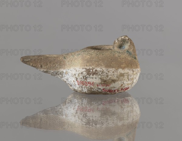 Oil lamp, 12th century, Iran. Ceramic utility oil lamp with a round container with a flattened bottom, a strongly elongated and flatly cut burner, and a rounded handle. Upper part covered with green glaze. Height 4.4 cm; width 9.8 cm; depth 6.0 cm. Persian; Islamic art; everyday life in the Middle East., An aged vessel shaped like a shallow, elongated bowl with a pointed spout and a rounded handle is shown resting on a reflective surface; the object has a rough, weathered texture with areas of brown, beige, white, greenish patina, and small blue-green spots, and its reflection appears below it against a neutral gray background.